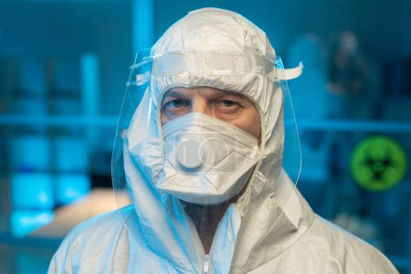 Téléchargez les photos : Face of male researcher in biohazard suit, respirator and protective screen looking at camera against interior of laboratory - en image libre de droit
