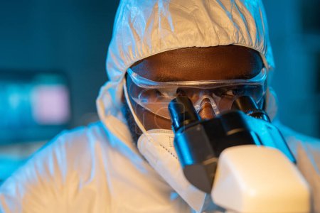 Téléchargez les photos : Close-up of young African American scientist in protective coveralls, respirator and eyeglasses looking in microscope in laboratory - en image libre de droit