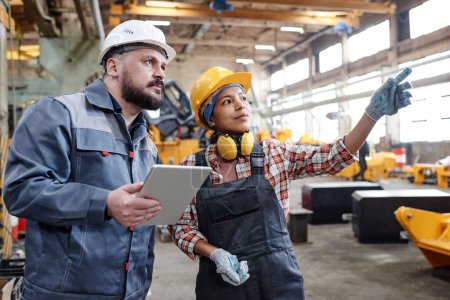 Foto de Young Hispanic female builder in workwear pointing at new equipment while discussing its characteristics with male colleague - Imagen libre de derechos