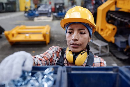 Foto de Young serious Hispanic female factory worker in gloves, hardhat and overalls taking pile of steel bolts out of plastic container - Imagen libre de derechos