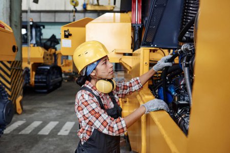 Foto de Young Hispanic female in protective helmet, coveralls and gloves checking engine of huge industrial machine before repairing - Imagen libre de derechos