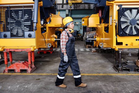 Foto de Young female technician in overalls, harhat and gloves moving along large industrial machines standing in rows in warehouse - Imagen libre de derechos