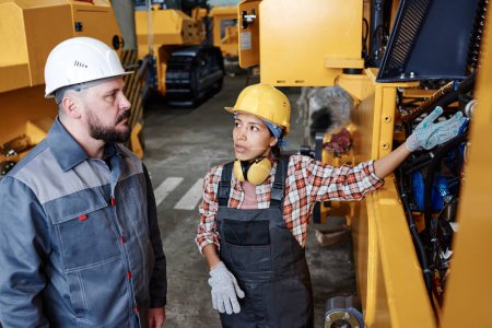 Foto de Young confident woman in workwear pointing at broken engine of industrial machine while consulting with her male colleague - Imagen libre de derechos