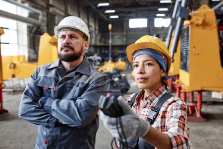 Foto de Two serious interracial workers of large factory looking at huge industrial machine while female technician regulating its height - Imagen libre de derechos