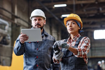 Téléchargez les photos : Two contemporary engineers with tablet and remote control looking at huge industrial machine while female regulating its work - en image libre de droit