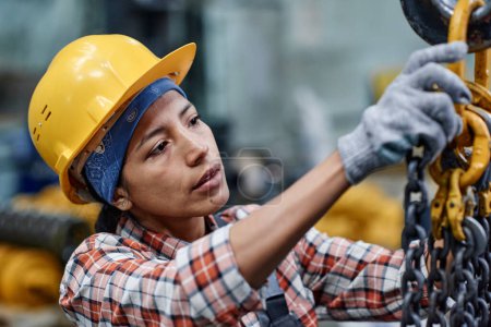Foto de Young Hispanic female engineer in hardhat and gloves checking chains of modern industrial machine while working in warehouse - Imagen libre de derechos