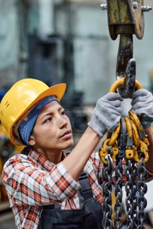 Foto de Young contemporary female engineer hanging bunch of metallic chains on hook of crane or other modern industrial machine - Imagen libre de derechos