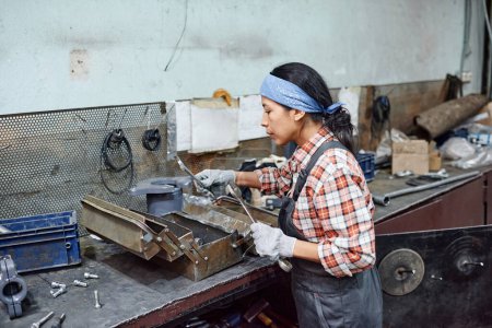 Foto de Young female worker of industrial plant choosing huge wrench for repair work while standing by workbench with metallic toolbox - Imagen libre de derechos