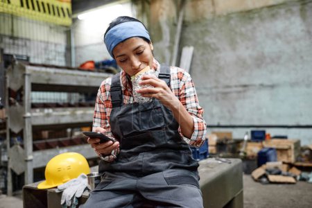 Foto de Young hungry Hispanic female engineer in coveralls and blue headband biting appetizing sandwich while having lunch at break - Imagen libre de derechos