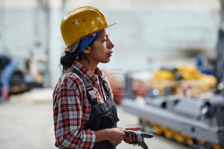 Foto de Side view of young tired female worker of factory in hardhat and coveralls standing in front of camera in large warehouse - Imagen libre de derechos