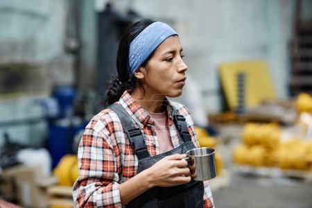 Foto de Young serious female builder in coveralls and blue headband holding mug with tea while having break in warehouse or workshop - Imagen libre de derechos