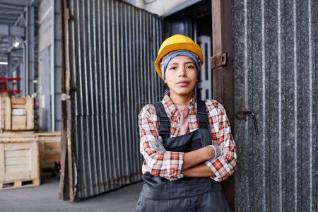 Foto de Young confident Hispanic female engineer in coveralls and protective helmet crossing arms by chest while standing in warehouse - Imagen libre de derechos
