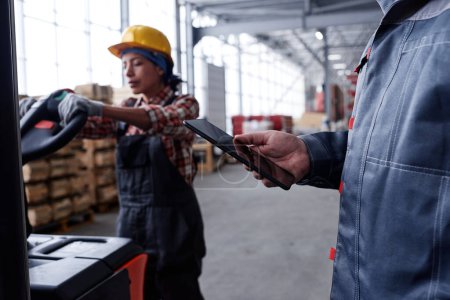 Foto de Hand of male manager of quality control in workwear holding tablet against young woman pushing cart with packages - Imagen libre de derechos