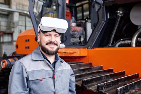 Foto de Happy mature worker of factory wearing vr headset and workwear looking at cameras while standing by industrial machine - Imagen libre de derechos