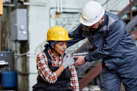 Foto de Foreman in hardhat and workwear standing by female engineer with contusion touching her shoulder against machine - Imagen libre de derechos