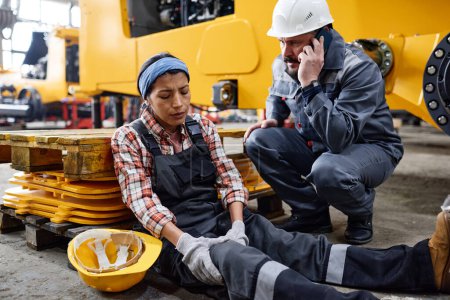 Photo for Young female engineer with hurting knee sitting on the floor by anxious male worker in safety helmet and uniform calling ambulance - Royalty Free Image