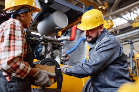 Foto de Mature workman in hardhat and workwear fixing shaft of industrial machine while his female colleague holding box with handtools - Imagen libre de derechos
