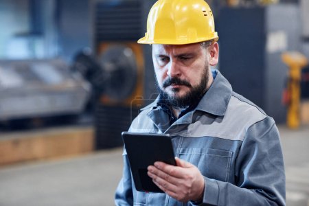 Téléchargez les photos : Mature bearded male worker of industrial plant in boilersuit and hardhat looking through data on screen of digital tablet - en image libre de droit