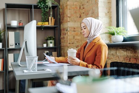 Téléchargez les photos : Young serious female accountant in hijab looking through financial papers and analyzing information about company finances - en image libre de droit