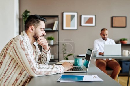 Foto de Serious bearded businessman in casual shirt planning work in front of laptop while sitting by desk against male colleague - Imagen libre de derechos