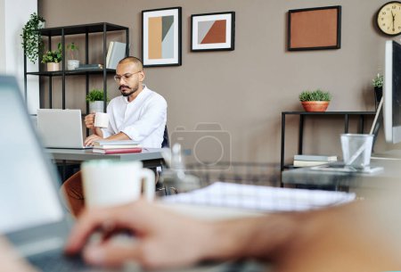 Téléchargez les photos : Serious young businessman with mug of tea looking through online data on laptop screen while analyzing it and having drink in office - en image libre de droit