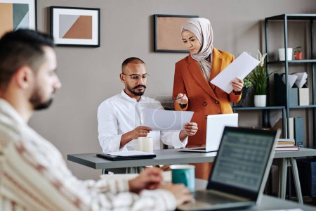 Téléchargez les photos : Young Muslim female and male economists discussing financial papers while businesswoman pointing at one held by colleague - en image libre de droit