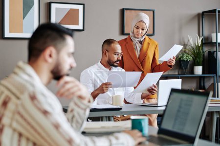 Foto de Two young Muslim coworkers looking through financial documents by workplace in front of male broker sitting in front of laptop - Imagen libre de derechos