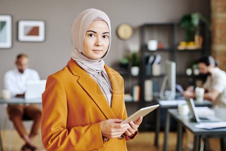 Téléchargez les photos : Young female business leader in hijab with tablet in hands looking at camera while networking against two male colleagues - en image libre de droit