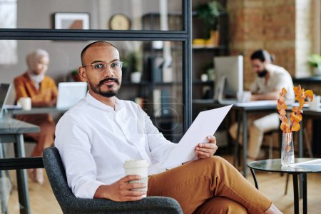 Foto de Young successful broker with glass of coffee and financial document sitting in armchair by small table against his coworkers - Imagen libre de derechos
