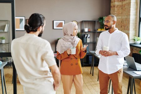 Photo for Group of young Muslim coworkers having coffee and discussing plans at break while standing in the center of openspace office - Royalty Free Image