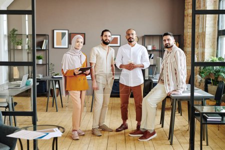 Téléchargez les photos : Group of four young successful employees in casualwear standing in the center of large modern openspace office and looking at camera - en image libre de droit