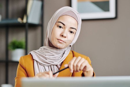 Photo for Young serious Muslim female in hijab sitting in front of laptop in office and looking at business partner on screen at digital meeting - Royalty Free Image
