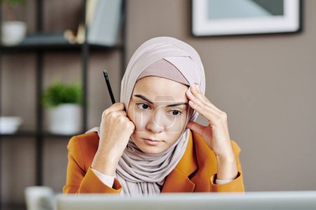 Téléchargez les photos : Serious young female employee in hijab looking attentively at laptop screen during digital meeting with business partner - en image libre de droit