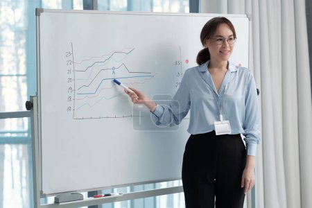 Photo for Happy young ceo of modern company making presentation of income increase to colleagues while pointing at graphs on board - Royalty Free Image