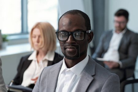 Téléchargez les photos : Young contemporary black man in formalwear and eyeglasses looking at camera while sitting against male and female colleagues - en image libre de droit
