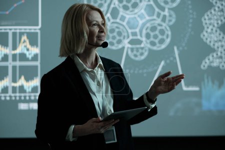 Photo for Happy blond mature businesswoman with tablet making speech at conference while standing against interactive board - Royalty Free Image