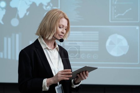 Photo for Serious female speaker or professor of economics with tablet reading lecture in auditorium while standing against interactive board - Royalty Free Image