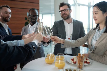 Téléchargez les photos : Happy interracial entrepreneurs clinking with flutes of champagne over table served with glasses of juice, canape and other snacks - en image libre de droit