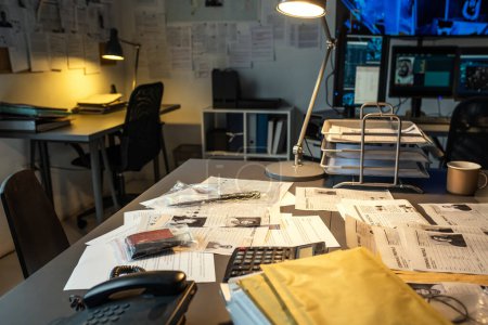 Téléchargez les photos : Part of workplace of modern fbi agent with documents, evidences, calculator and telephone on desk lit by lamp in small office - en image libre de droit