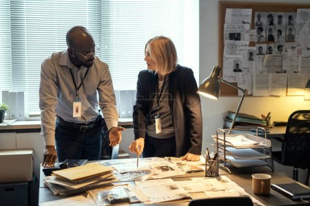 Photo for Two interracial agents of FBI discussing criminal profiles at meeting in bureau while one of them pointing at photo of suspect - Royalty Free Image