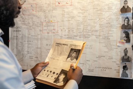 Foto de Young African American male FBI agent looking through criminal profile of suspect in front of board with map and photos of suspects - Imagen libre de derechos