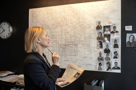 Mature female agent of FBI with documents matching facts of crime series while standing in front of board and looking at map