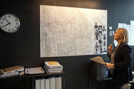 Foto de Mature blond serious female investigator looking at map on board while thinking of ideas about possible location of serial killer - Imagen libre de derechos