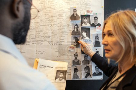 Foto de Blond confident female detective pointing at photo of suspect on board over map while matching facts of serial crimes with colleague - Imagen libre de derechos