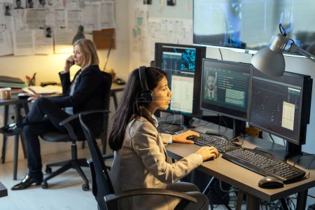 Foto de Serious female agent of intelligence service sitting in front of computer monitors in office and processing personal data of criminals - Imagen libre de derechos