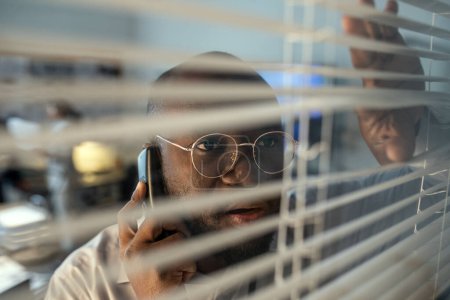 Téléchargez les photos : African American male security system department staff talking on the phone while looking through venetian blinds in office - en image libre de droit