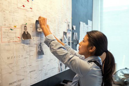 Foto de Young Hispanic female agent of FBI in uniform pinning photos of several male suspects on map while suggesting their location - Imagen libre de derechos