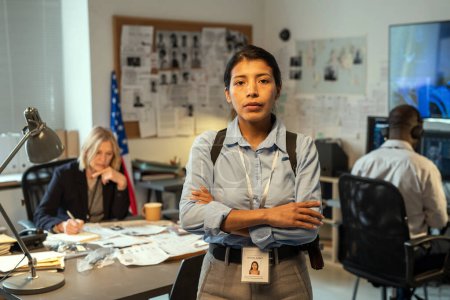 Téléchargez les photos : Young Hispanic female leader of intelligence service or police department in uniform crossing arms on chest while standing against coworkers - en image libre de droit