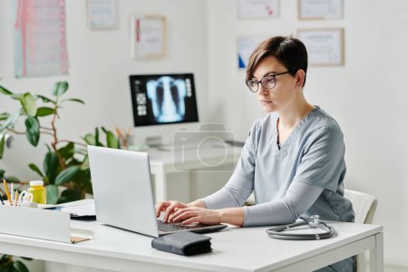Téléchargez les photos : Young serious female doctor in medical scrubs typing on laptop keyboard while consulting online patient by workplace in office - en image libre de droit