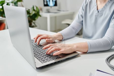 Foto de Manos de médico joven u otro especialista en escribir uniforme azul en el teclado del ordenador portátil mientras está sentado en el lugar de trabajo - Imagen libre de derechos